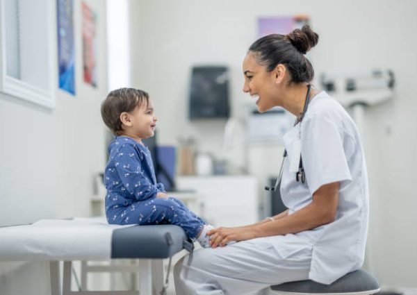 A sweet little toddler sits up on an exam table in a doctors office as her female doctor conducts a routine check-up.  The little girl is dressed casually and appears happy as her doctor engages with her.